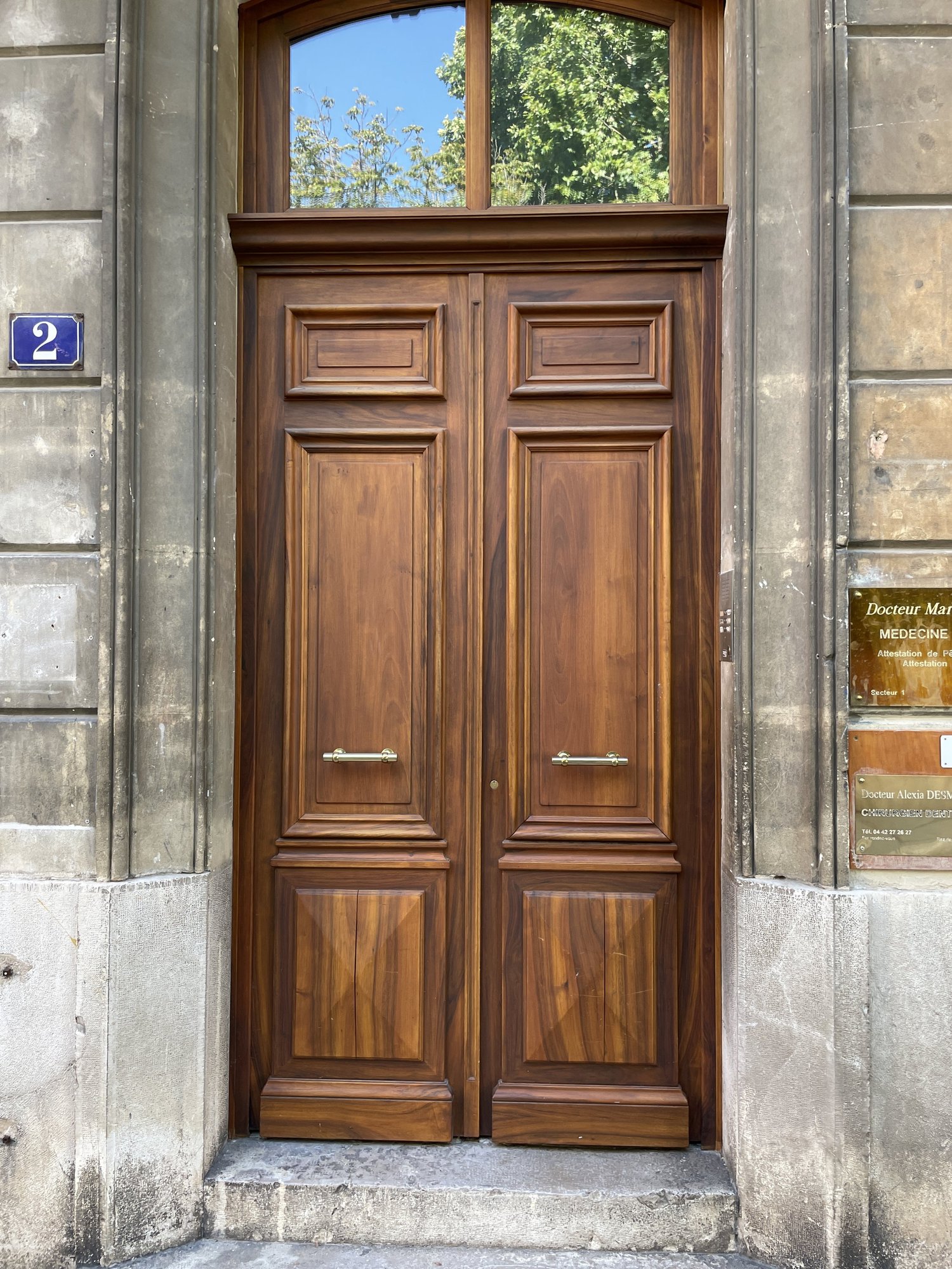 Porte en bois massif avec deux panneaux, une fenêtre en arc en haut, et des poignées en métal, devant un mur en pierre.