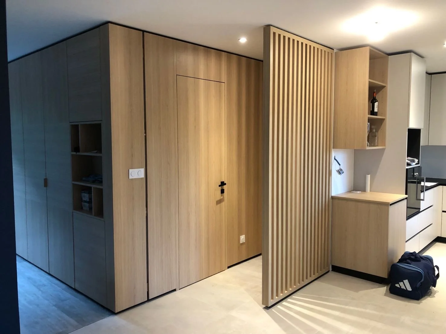 Une pièce moderne avec des meubles en bois clair, comprenant des armoires, des étagères, un panneau séparateur en bois ajouré et une petite unité de rangement, avec un sac de sport noir sur le sol.