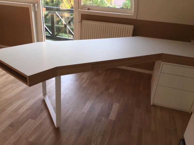 Un bureau en angle avec un dessus blanc et un meuble à tiroirs blanc, situé près d'une fenêtre laissant entrer la lumière naturelle.