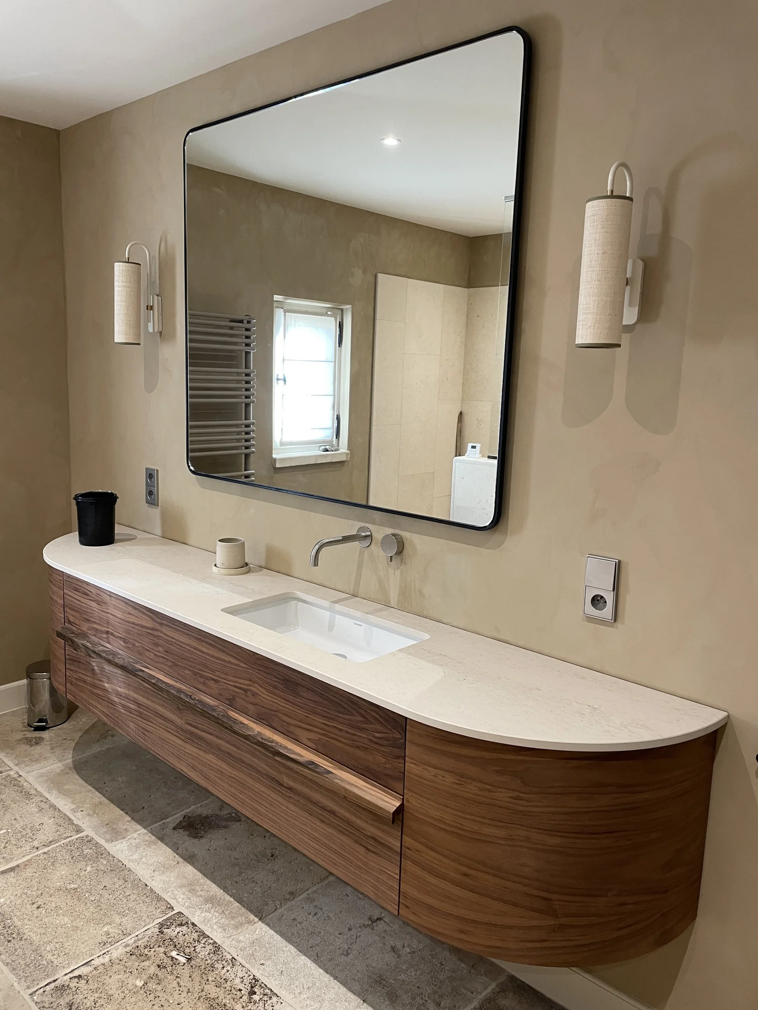 Grand miroir rond au-dessus d'un meuble en bois avec un lavabo intégré, éclairé par deux appliques murales, dans une salle de bain moderne avec murs beige et sol en pierre.
