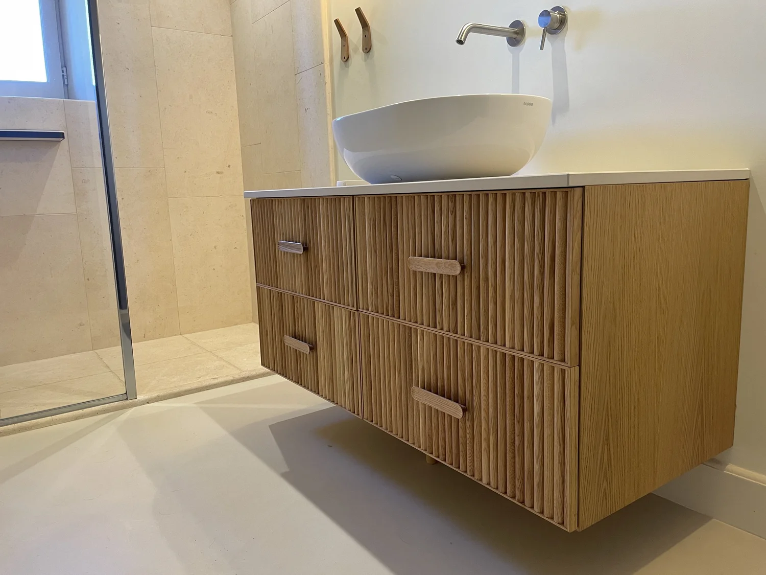 Meuble de salle de bain en bois clair avec tiroirs à poignées en bois, lavabo rond en céramique blanche, et deux robinets au mur. Salle de bain avec mur beige et sol clair. Fenêtre à gauche.