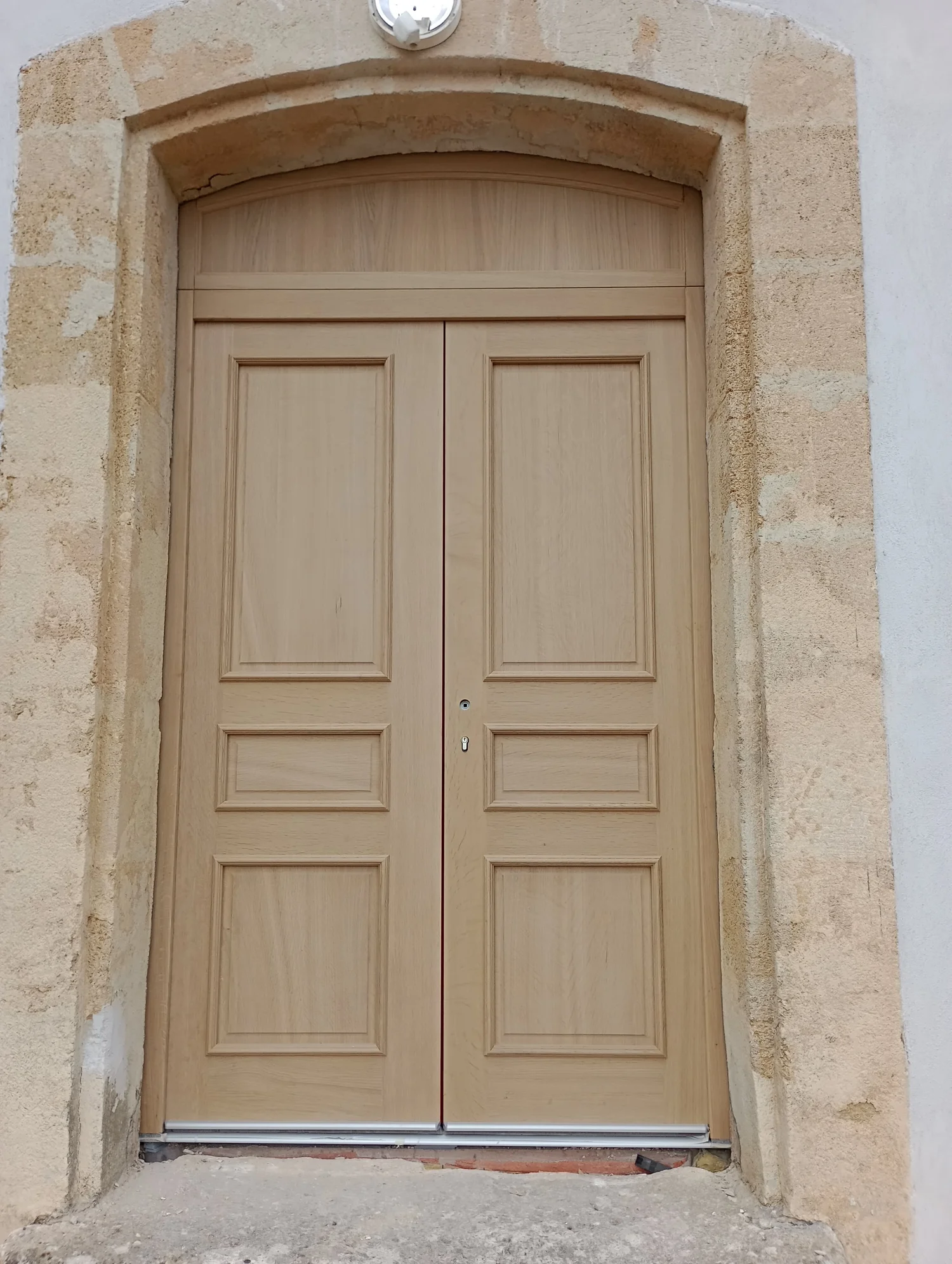 Porte en bois double avec quatre panneaux chacun, encadrée par un mur en pierre et un cadre en pierre biseauté, en haut un arc en pierre. 