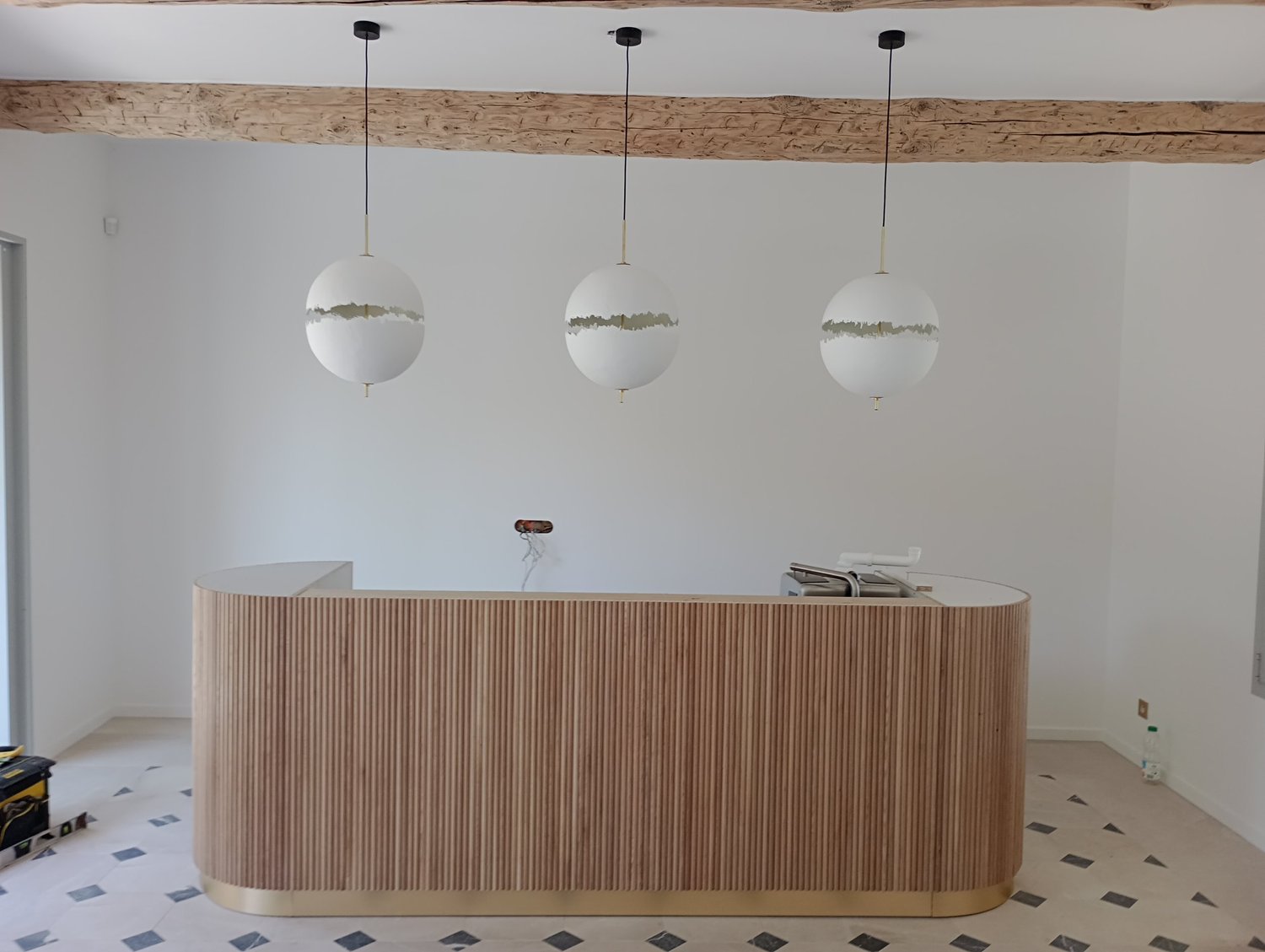 Un comptoir en bois clair courbé dans une pièce en cours de rénovation, avec trois lampes suspendues au plafond et un sol en carreaux blanc et noir.
