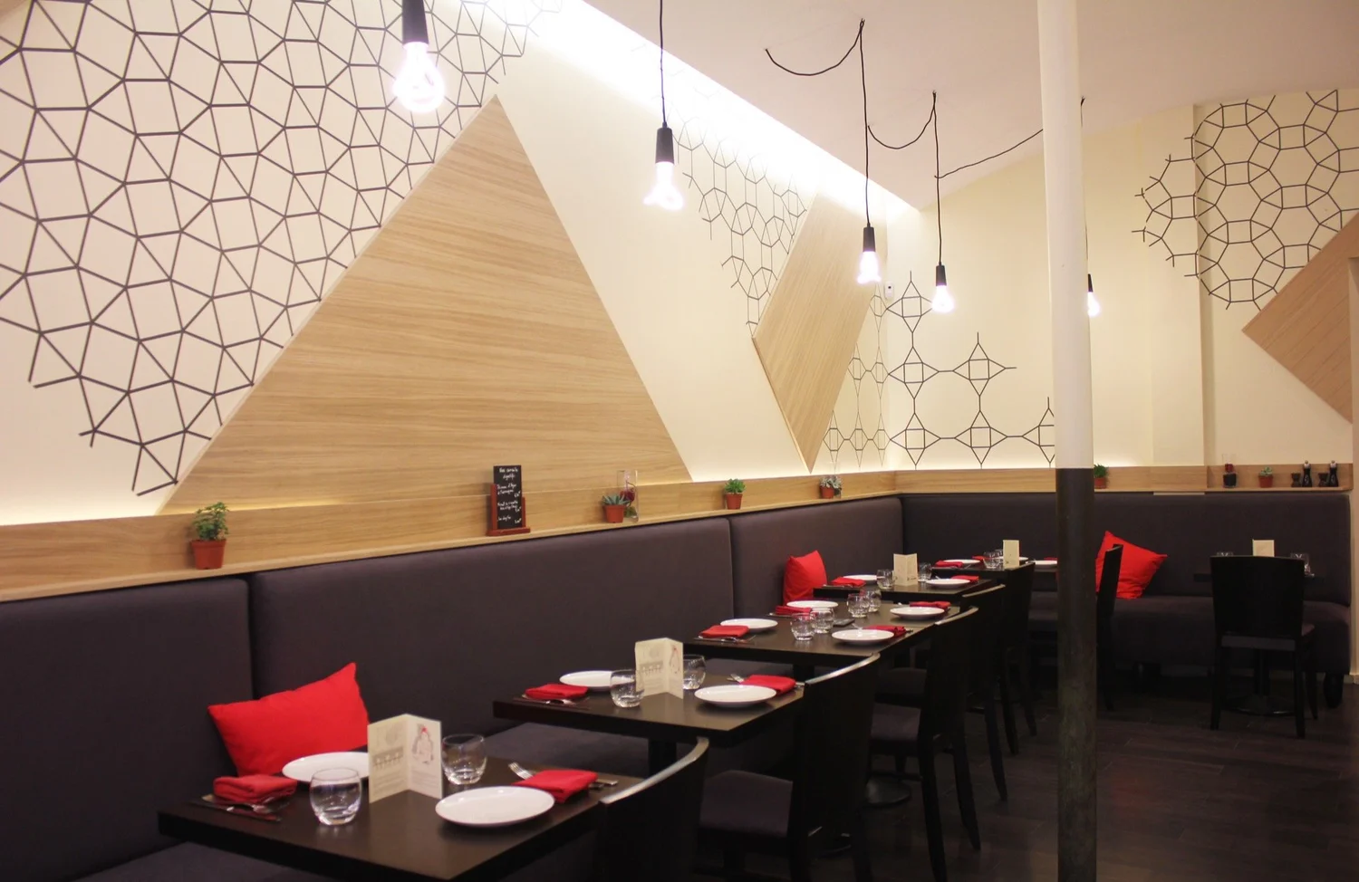 Salle à manger dans un restaurant moderne, avec des murs décorés de motifs géométriques, des tables avec assiettes, verres et serviettes rouges, des chaises noires, et des coussins rouges sur un banc noir.